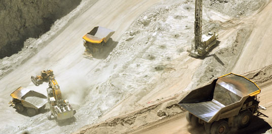 Ein Tagebau in dem Kies oder ähnliches Material gefördet wird. Im Vordergrund ist ein leere Kipplader, der gerade auf einem Weg in den Tagebau hinuter fährt. Im Hintergrund werden andere Kipplader durch einen Bagger beladen.